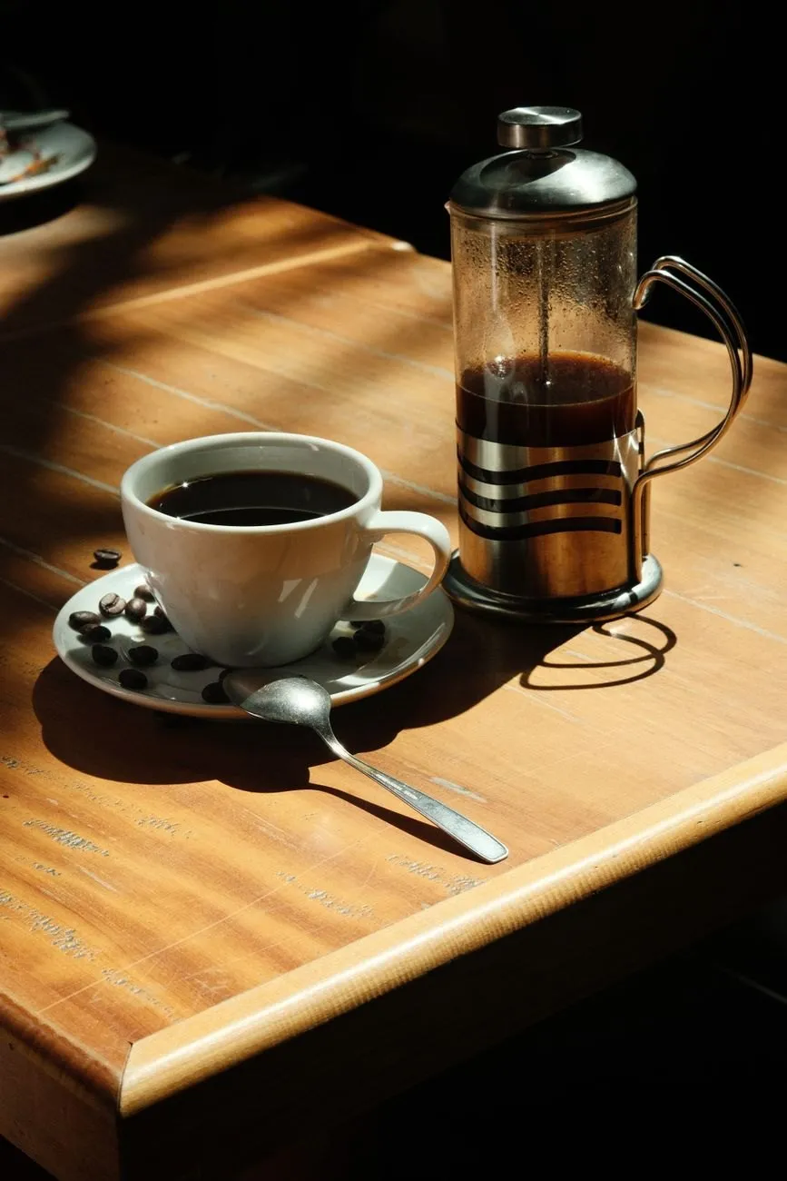 A cafetière of coffee next to a white cup and saucer on a sunlit wooden table