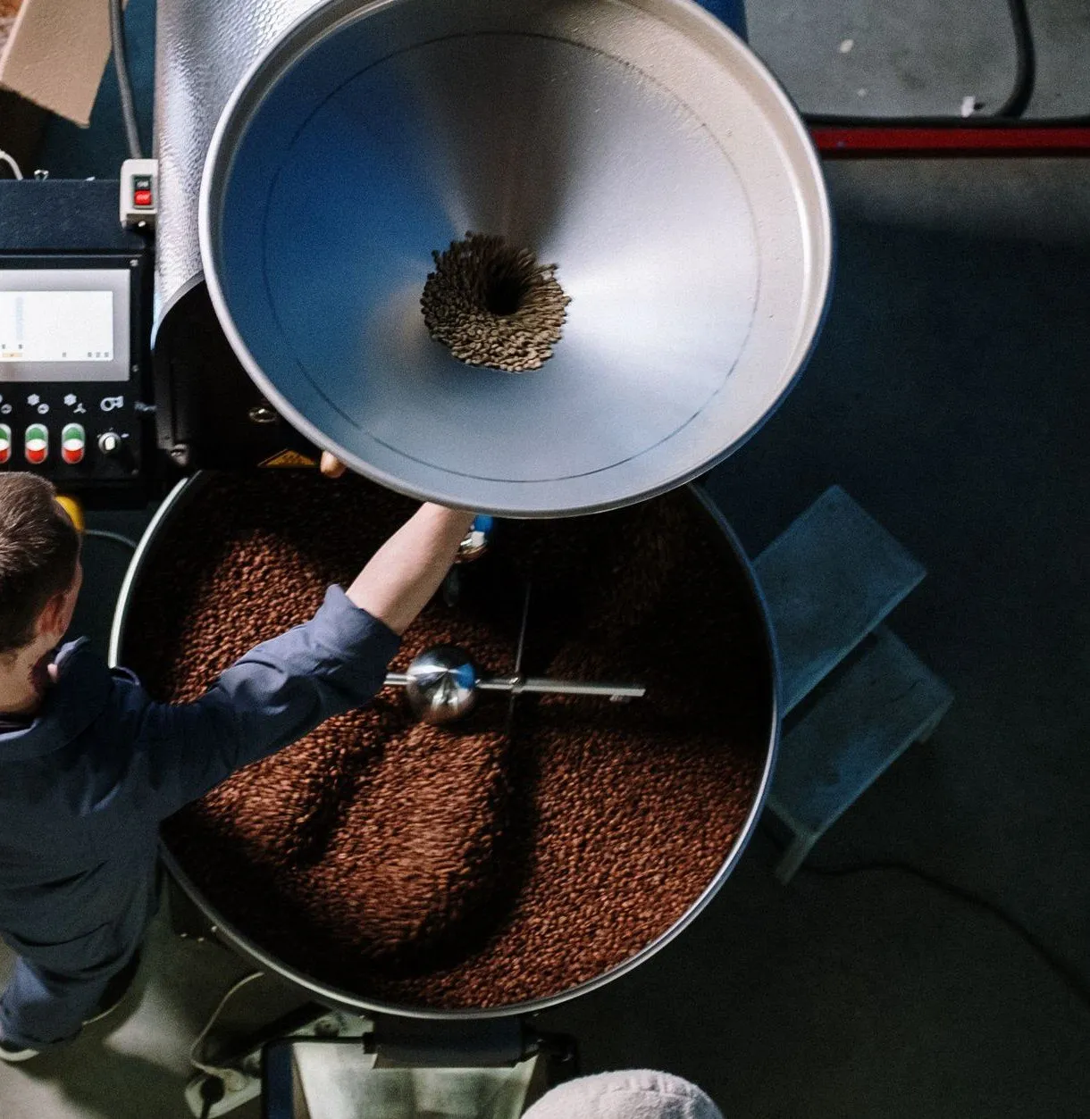 The roaster loading green coffee into the drum hopper, seen from the mezzanine above
